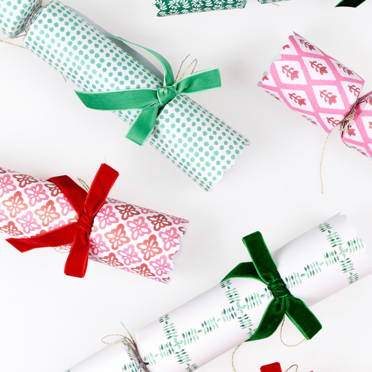 Four Christmas cracker ornaments wrapped in patterned paper and tied with satin ribbon bows in green, red, and pink, arranged on a white background.