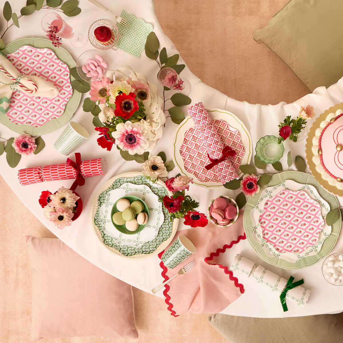 A styled tabletop flat-lay of a coordinated table setting with pink and green patterned plates, pink napkins with red scalloped edges, floral centerpieces, wrapped silverware, and decorative greenery on a beige surface.