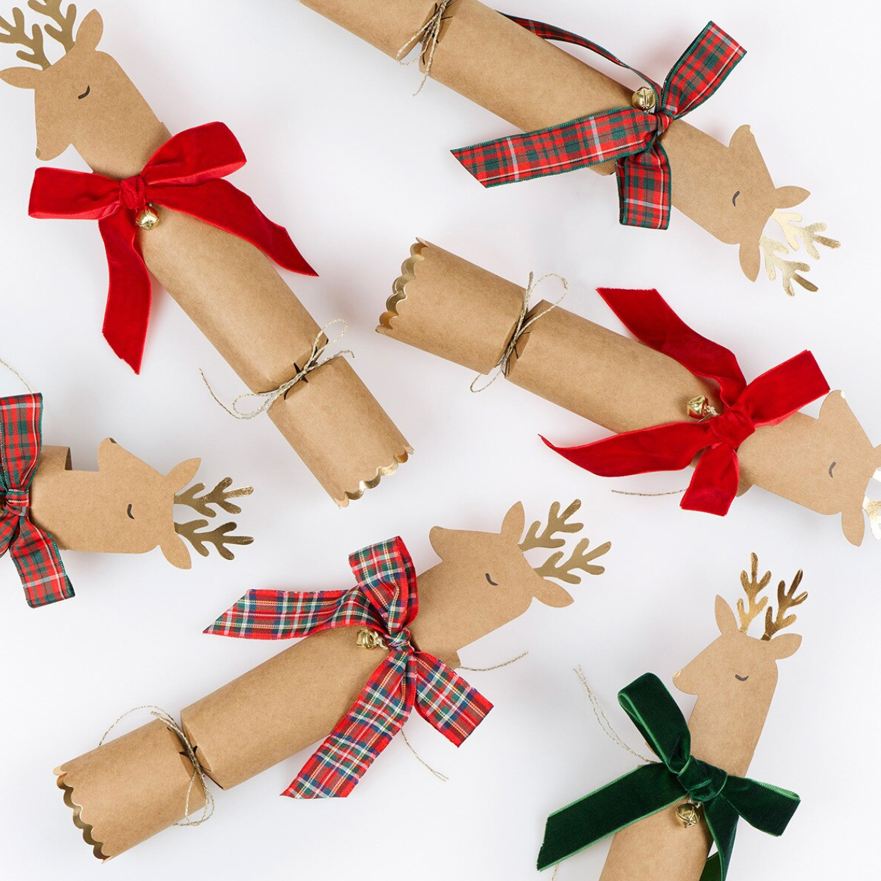 Five handmade paper cracker Christmas ornaments shaped like reindeer, wrapped in brown kraft paper with small wooden eyes and noses, decorated with red, green plaid, and dark green velvet bows, and gold jingle bells, arranged on a white background.