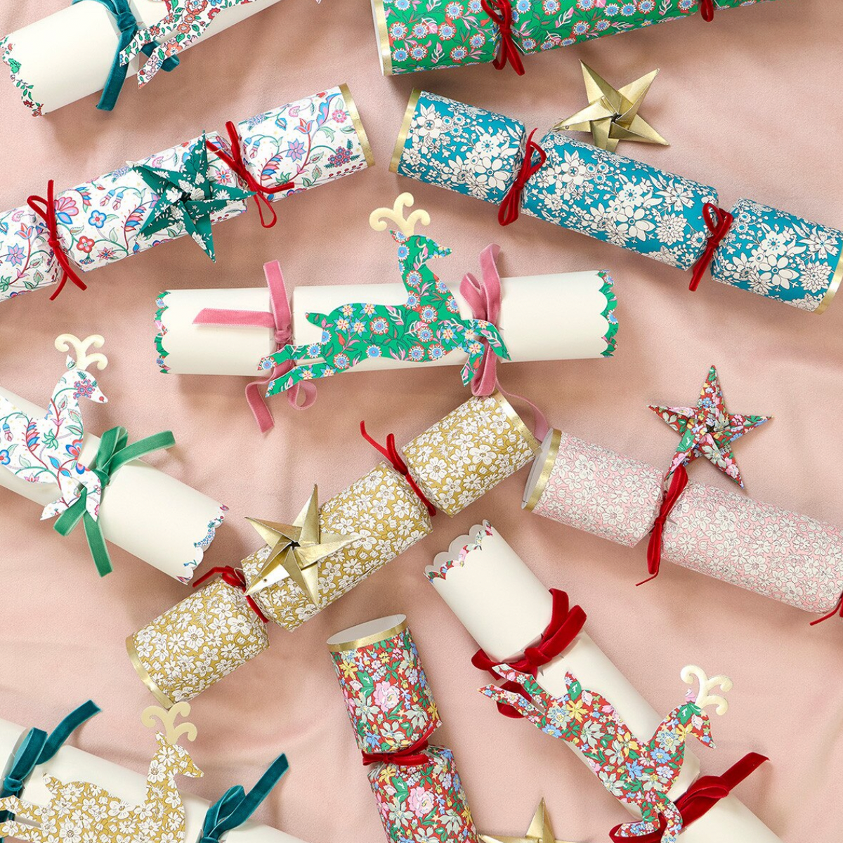 A set of Christmas cracker ornaments arranged on a soft pink background. Each cracker is wrapped in festive floral and patterned paper, tied with ribbon, and decorated with a small reindeer or star ornament.