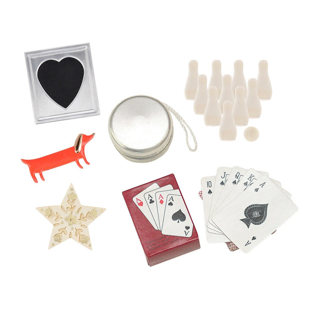 A flat-lay collection of small decorative and game items on a white background, including a heart-shaped black-and-silver ornament, a red dachshund figure, a gold star, a round silver tin, a red card deck, playing cards, and several small beige bottle-like pieces.