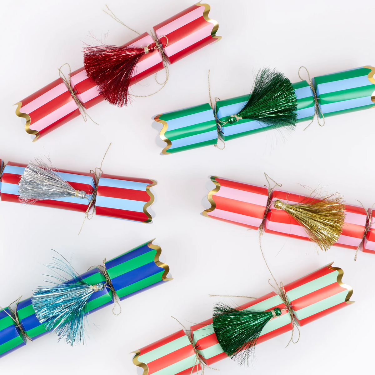 Six festive paper cracker ornaments arranged on a white background, each decorated with colorful striped bands and a metallic tassel. The cracker designs include red and white, green and blue, and red and mint green, with tassels in red, green, silver, blue, gold, and green.