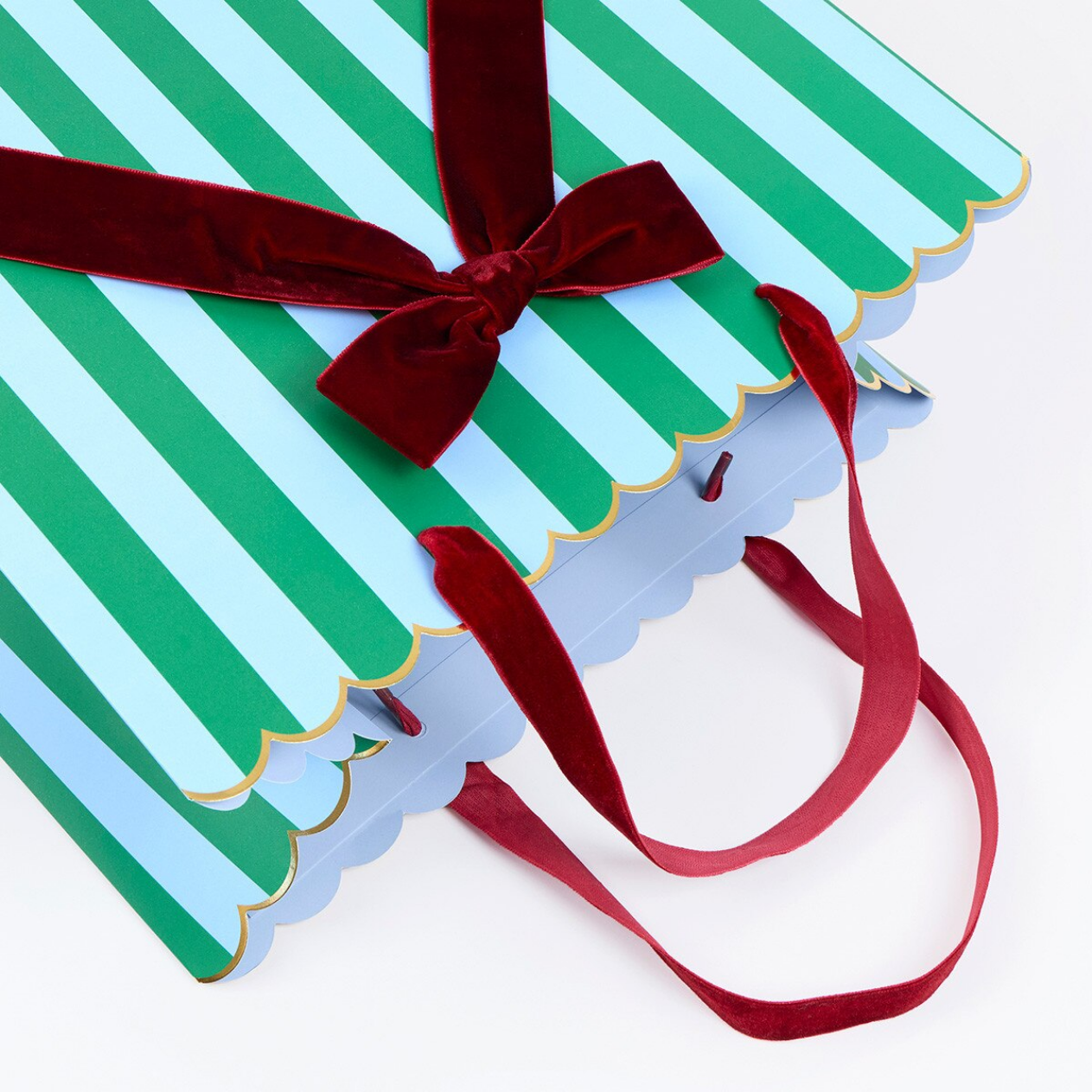 A close-up of a striped gift bag or packaging with alternating green and light blue vertical stripes, scalloped edges, a dark red velvet bow, and long red ribbon handles on a white background.