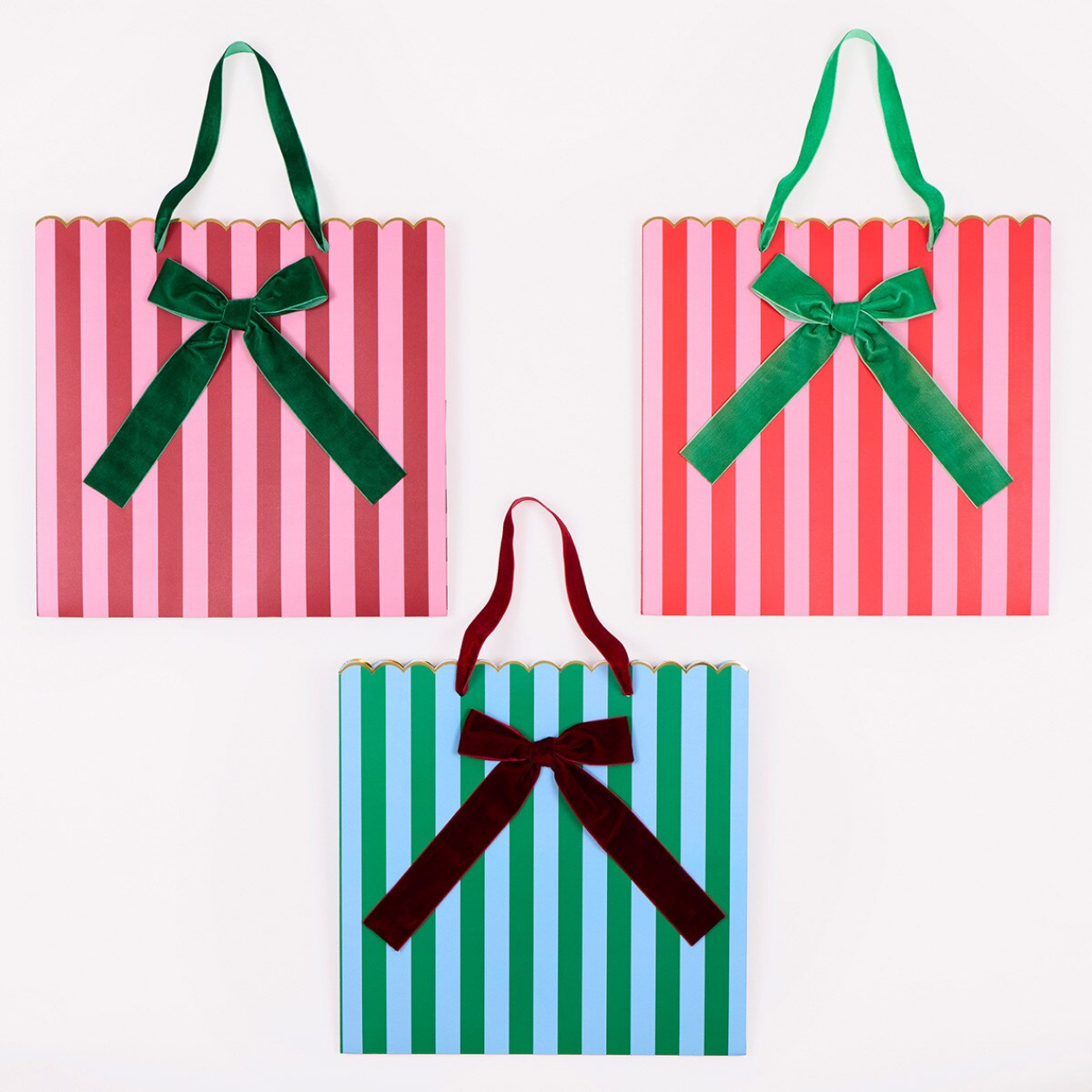 Three striped gift bags arranged on a white background: two pink-and-red striped bags with green velvet handles and bows, and one blue-and-green striped bag with a dark burgundy velvet handle and bow.