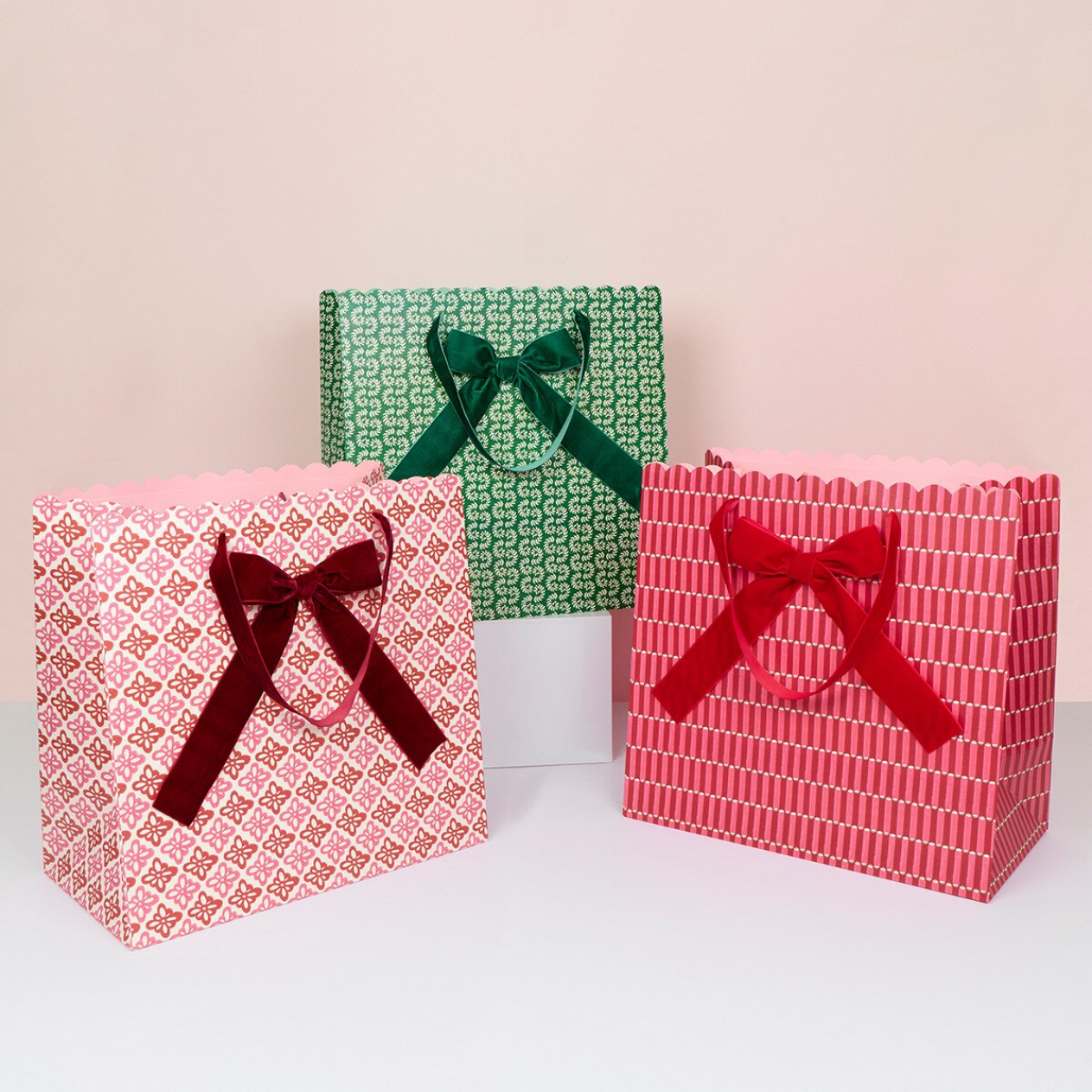 Three gift bags with patterned wrapping paper and ribbon handles, arranged on a white surface against a pale pink background. One bag is pink with a red geometric pattern and a dark red ribbon, one is green with a white geometric pattern and a dark green ribbon, and one is red with a white vertical pattern and a bright red ribbon.
