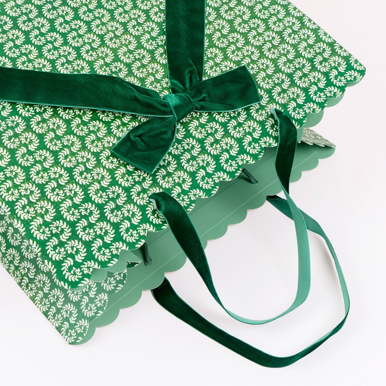 A green gift bag with a repeating white floral pattern, scalloped edges, and dark green velvet ribbon handles tied in a bow, photographed on a white background.