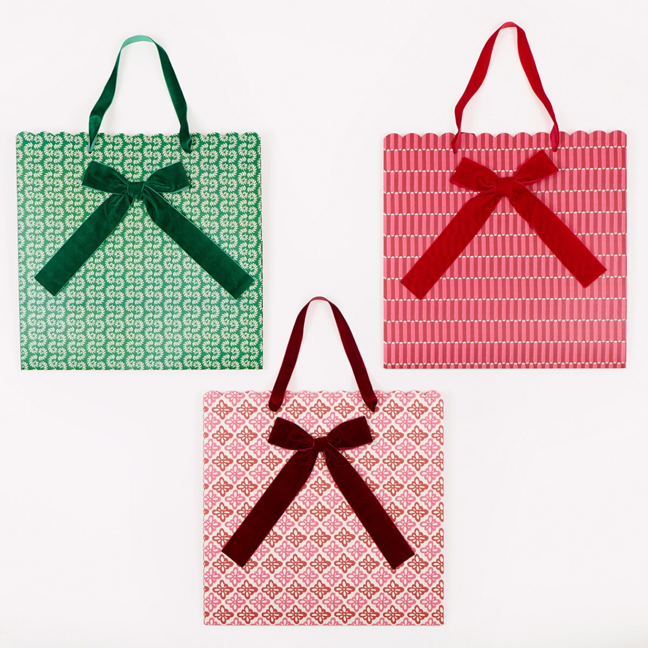 Three patterned gift bags arranged on a white background: one green bag with a green bow, one pink bag with a red bow, and one pink bag with a dark burgundy bow. Each bag has a repeating geometric pattern and a large ribbon handle.