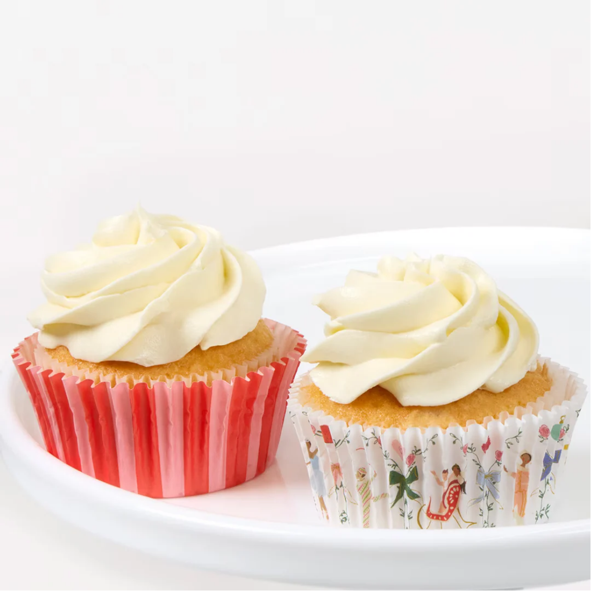 Two frosted cupcakes on a white plate against a clean white background. One cupcake has pale cream frosting in a pink striped paper liner, and the other has pale cream frosting in a white paper liner with a small multicolor floral and figure print.
