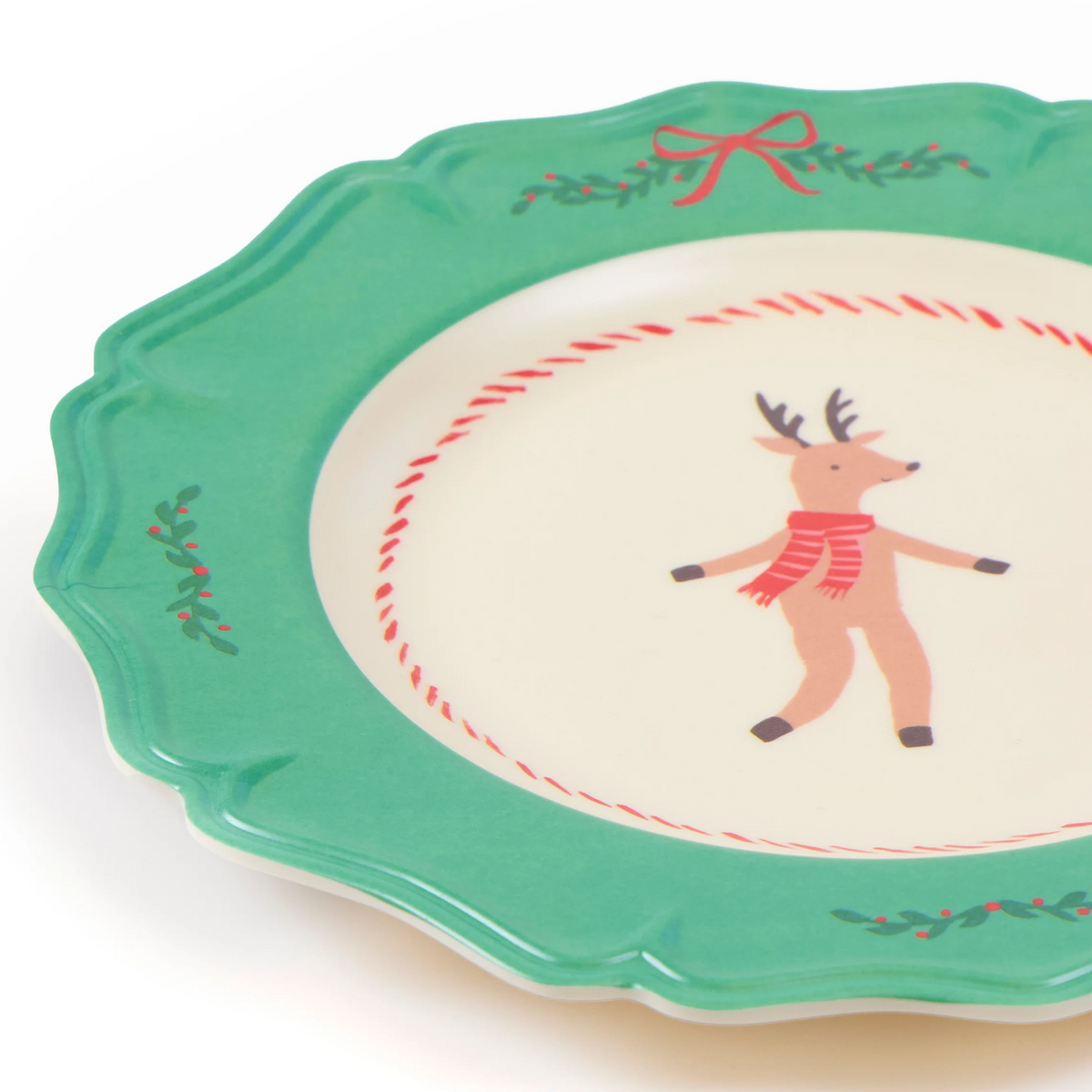 A close-up of a ceramic plate with a scalloped green rim, decorated with a red bow, holly leaves, and a small reindeer wearing a red scarf in the center.