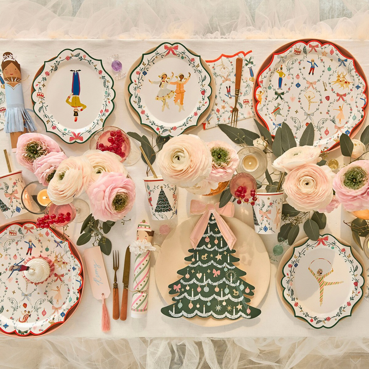 A styled tabletop display of decorative ceramic plates, pink flowers, candles, and small tableware with a whimsical holiday and ballet theme, arranged on a white surface with sheer tulle fabric.