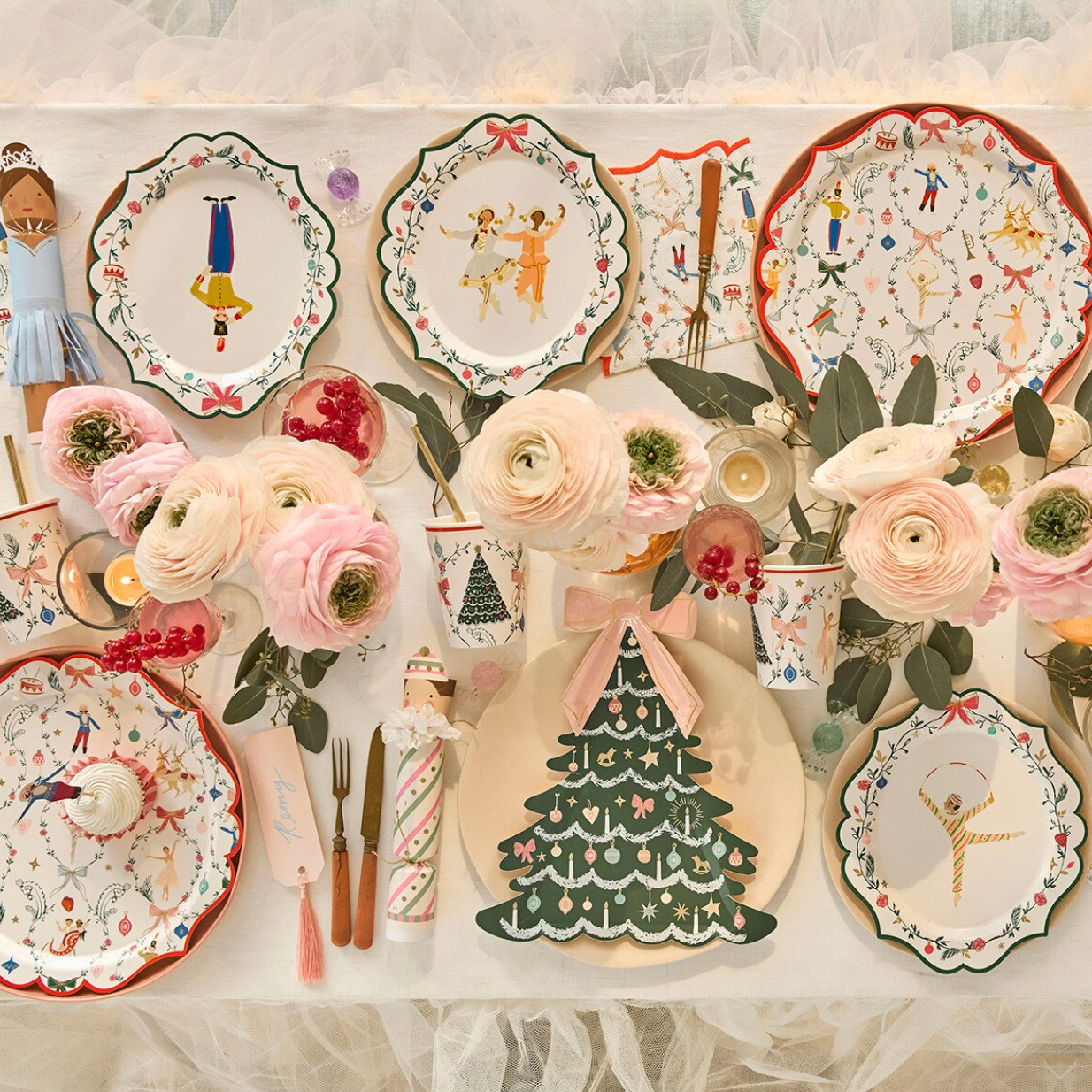 A styled tabletop display of decorative ceramic plates, pink flowers, candles, and small tableware with a whimsical holiday and ballet theme, arranged on a white surface with sheer tulle fabric.