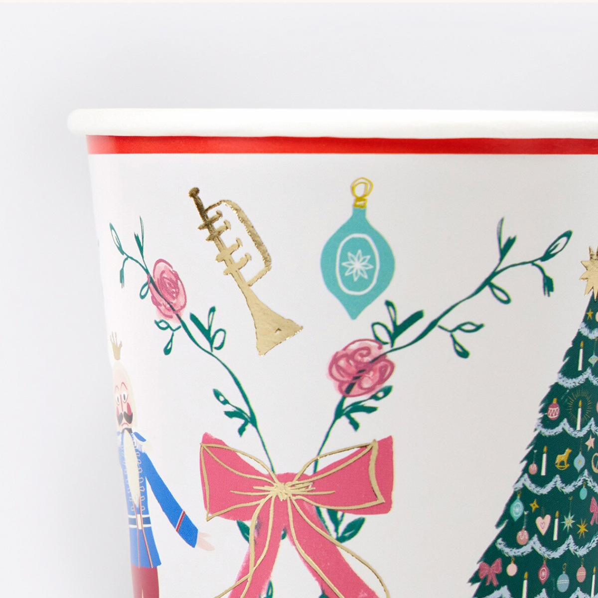 A close-up of a white paper cup printed with a festive Christmas design, featuring a pink bow, gold trumpet, teal ornament, pink flowers, a nutcracker, and part of a decorated Christmas tree.