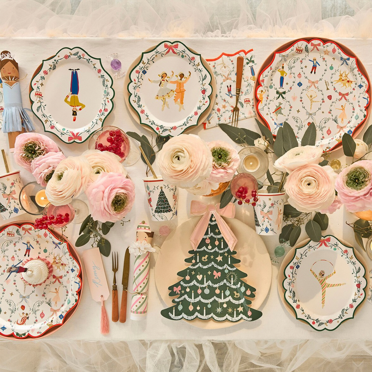 A styled tabletop arrangement of decorative ceramic plates, cups, napkins, and floral centerpieces in soft pink, green, red, and white, photographed from above on a white fabric backdrop.