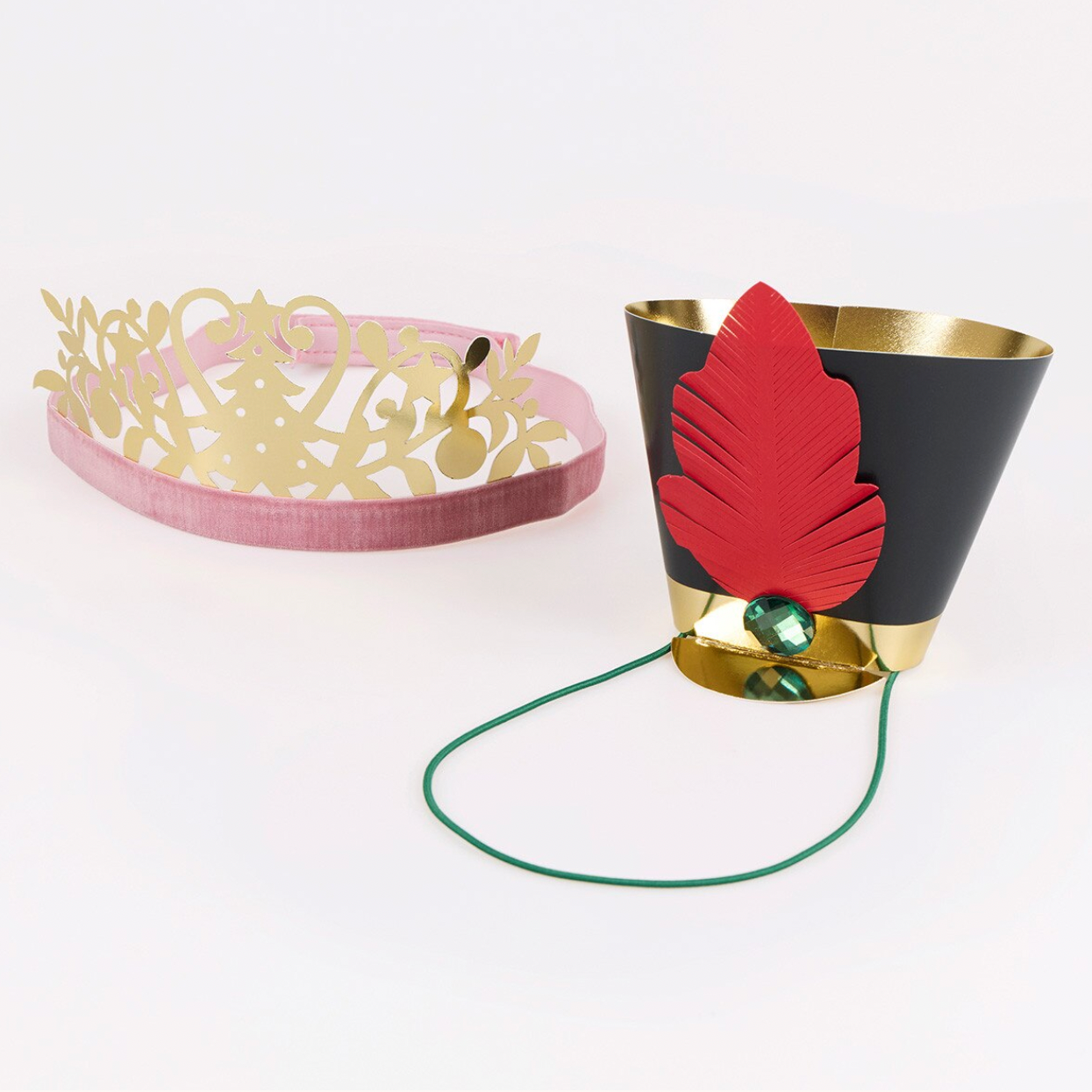 A toy costume accessory set on a white background, featuring a gold paper crown on a pink headband and a black paper hat with a red paper leaf, gold trim, a green hanging cord, and a green jewel.