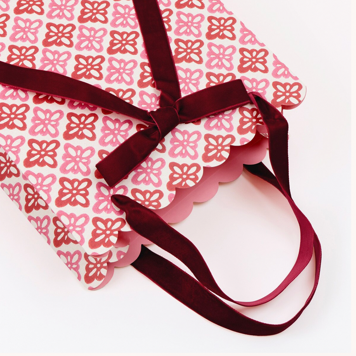 A tote bag with a pink and red floral pattern on white fabric, featuring a scalloped edge and dark burgundy velvet ribbon handles, laid flat on a white background.