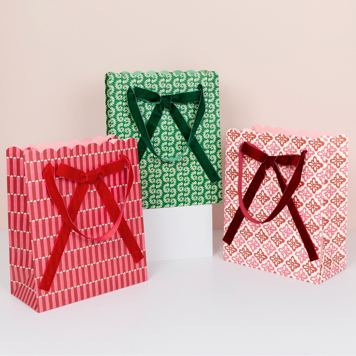 Three gift bags in pink and green holiday patterns with velvet ribbon handles, displayed on a white surface against a light beige background.