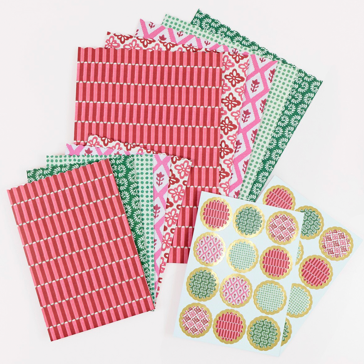 A set of patterned paper sheets and circular patterned stickers arranged on a plain white background. The papers feature various festive holiday motifs including stripes, florals, and geometric patterns in red, pink, green, and white, with some sheets showing gold accents.