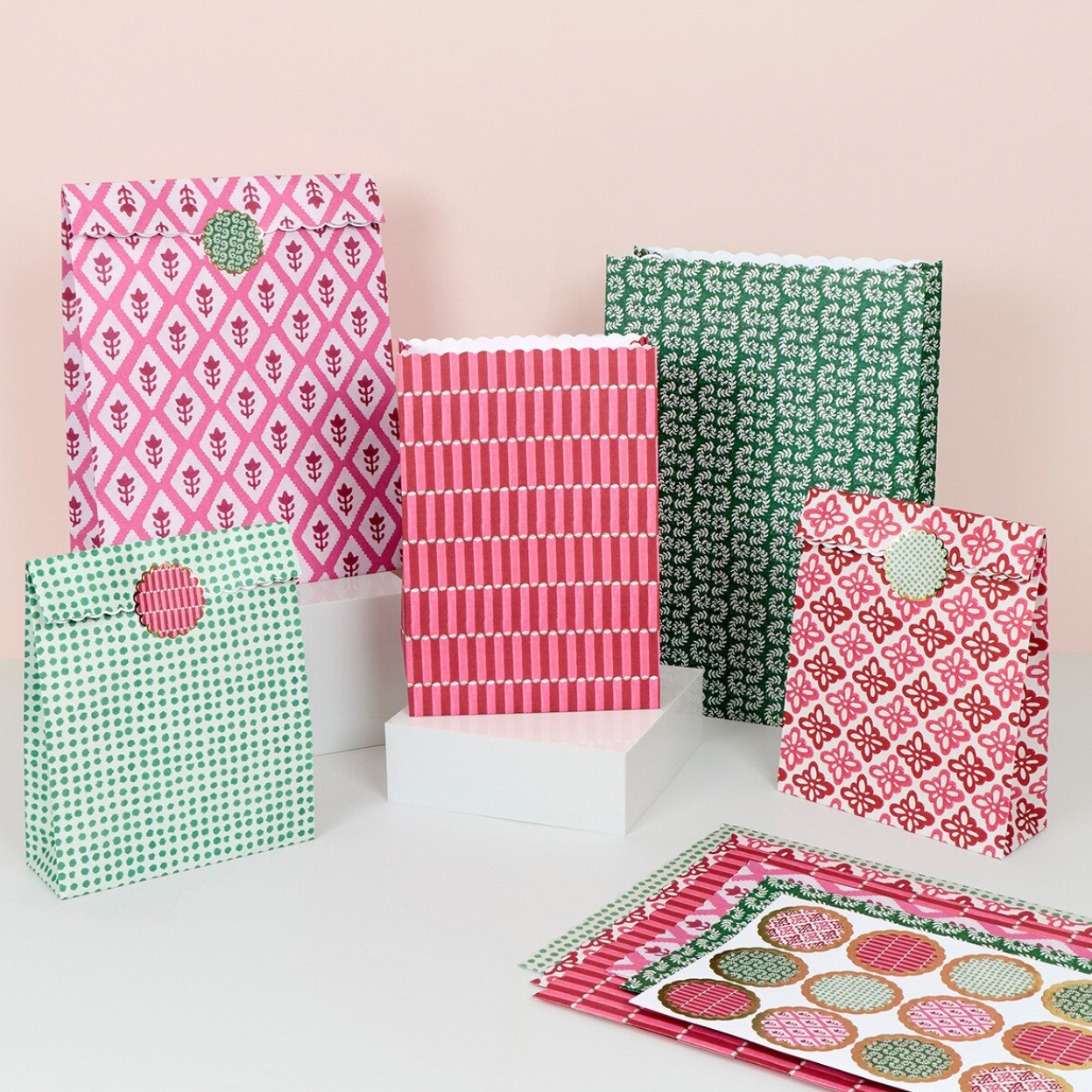 A set of patterned gift bags and matching paper goods packaging arranged on white blocks against a pale pink background. The bags feature various colorful prints including pink and green floral motifs, green and white dots, pink and white stripes, and pink and white geometric designs, with one bag showing a circular green patch on the front.