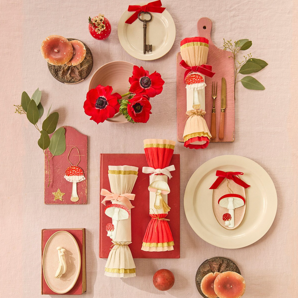 A styled tabletop flat lay of red and cream tableware and decorative accessories on a pale pink fabric background, including plates, rolled napkins tied with red ribbons, a key, mushrooms, flowers, and small wooden decor pieces.