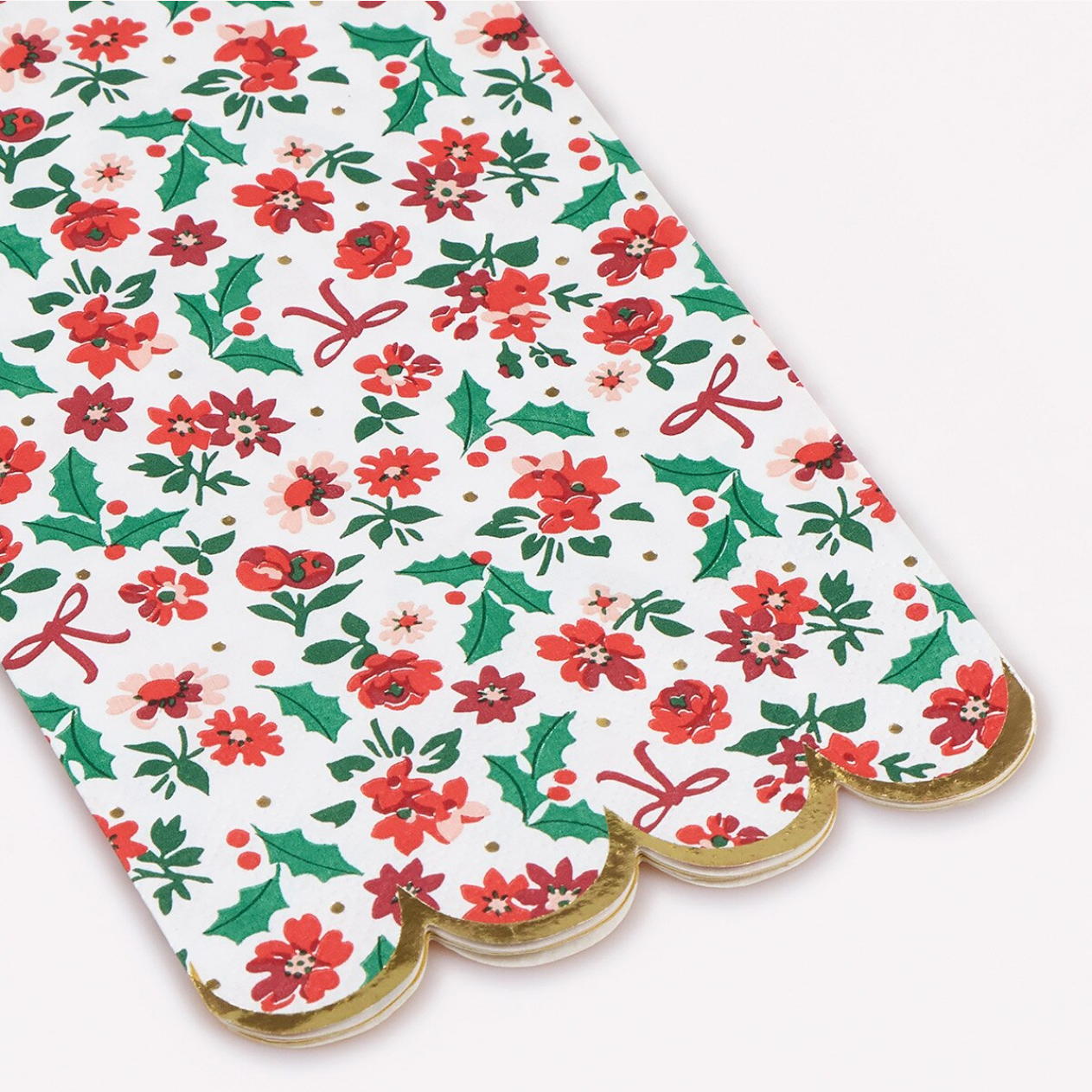 A stack of scalloped-edge paper napkins featuring a festive Christmas floral pattern with red poinsettias, green holly leaves, red bows, and small gold polka dots on a white background.