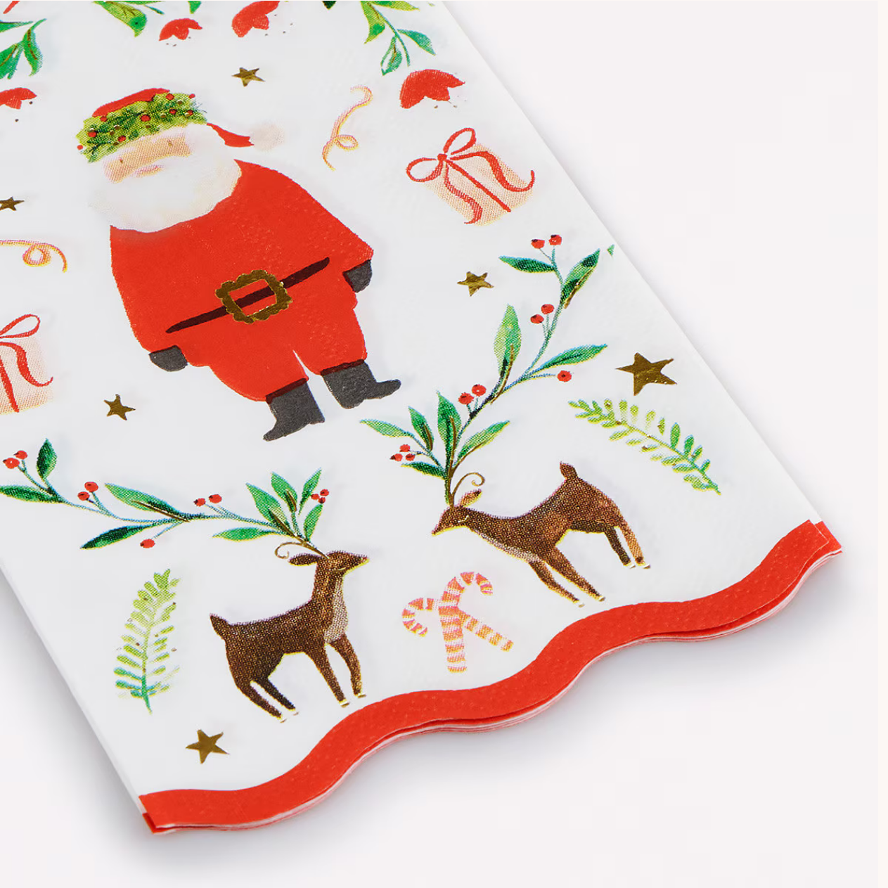 A close-up of a white holiday-themed napkin with a scalloped edge, printed with a Santa Claus figure, green holly and pine branches, brown deer, candy canes, and gold stars.