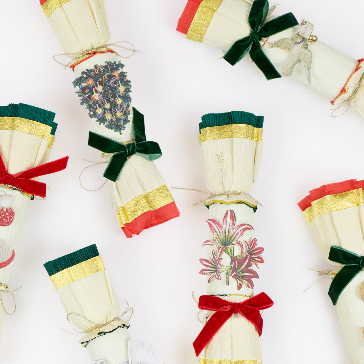 A styled flat-lay of multiple cream-colored Christmas cracker ornaments with gold trim, green and red ribbons, and floral or botanical prints, arranged on a clean white background.