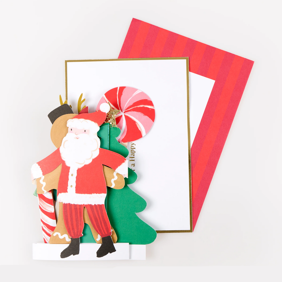 A festive layered paper craft or greeting card design featuring a cutout Santa Claus figure, a green Christmas tree, a red-and-white striped candy cane, and a striped red-and-white envelope behind a white card on a light background.