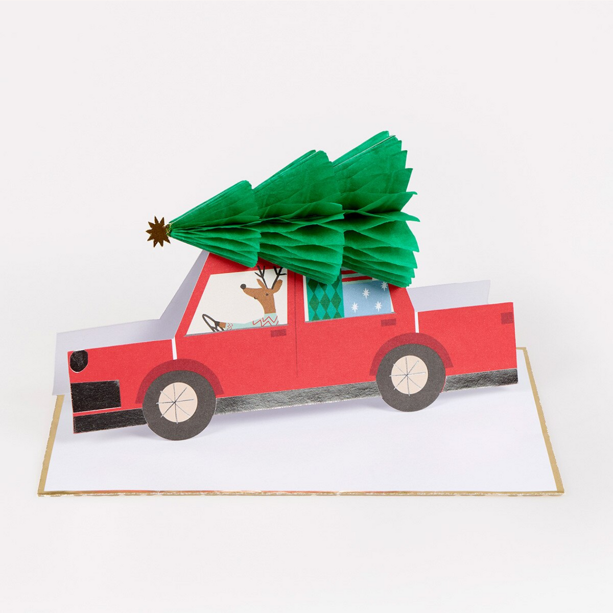 A 3D papercraft pop-up card featuring a red pickup truck carrying a green Christmas tree, with a reindeer visible in the driver's seat, set against a plain white background.