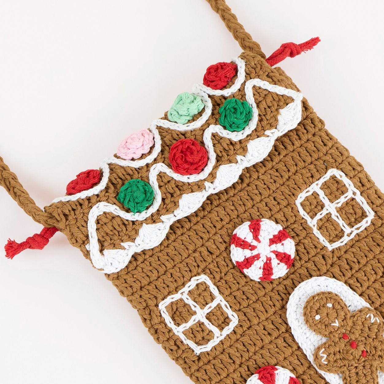 A crocheted gingerbread house shoulder bag in brown yarn with white icing details, red and green candy decorations, and a red-and-white peppermint candy motif, shown on a plain light background.