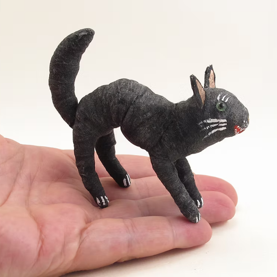 A small black cat figurine with white whiskers and a green eye, displayed on a person's hand against a plain white background.