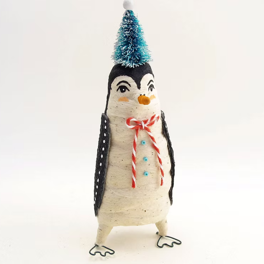 A handmade penguin figurine wrapped in textured cream-colored paper, with black wings, a small orange beak, a red and white striped bow around its neck, and a blue glittery Christmas tree hat on top, set against a plain white background.