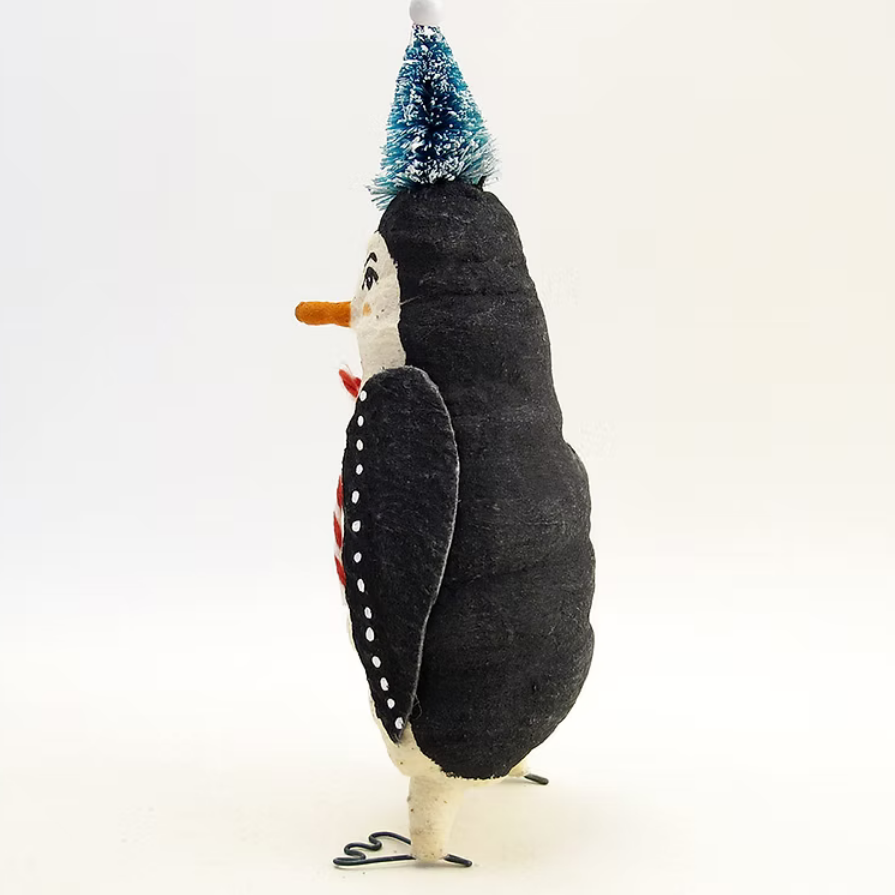 A handmade felt penguin ornament or decoration shown from the side on a white background. The penguin has a black felt body, white belly and beak, a small orange carrot nose, a red scarf with white dots, and a tiny blue-and-white glittery party hat.
