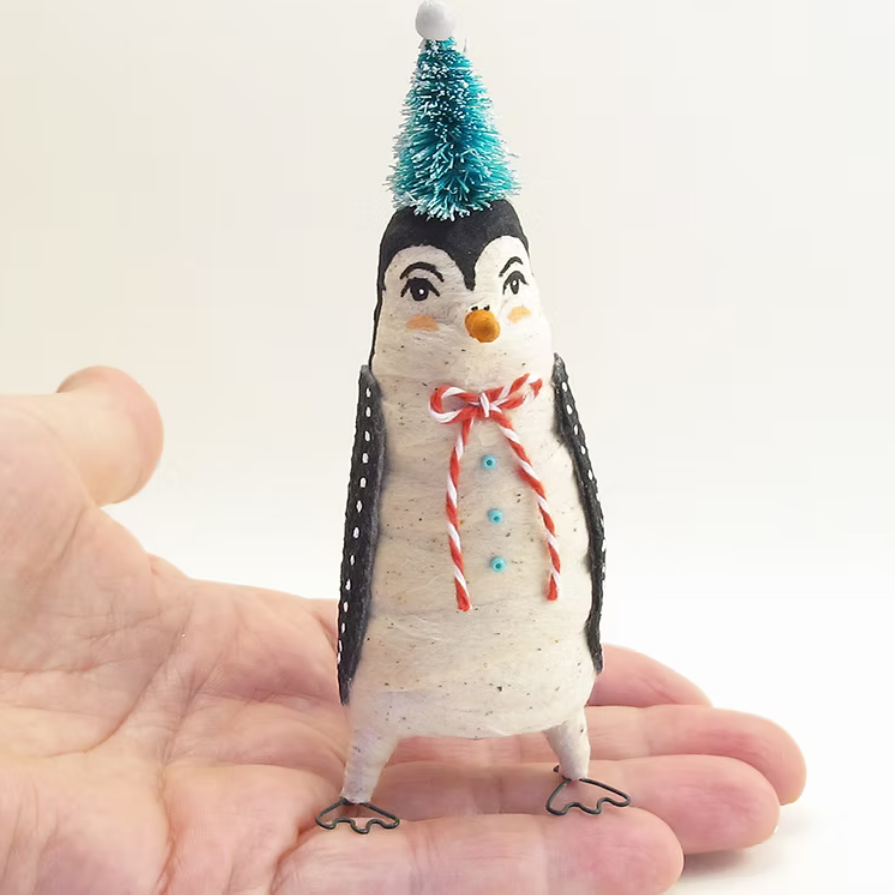 A small penguin-shaped decorative figure with black and white body panels, a turquoise party hat, and a red-and-white striped bow tie, standing on a person's open palm against a plain light background.