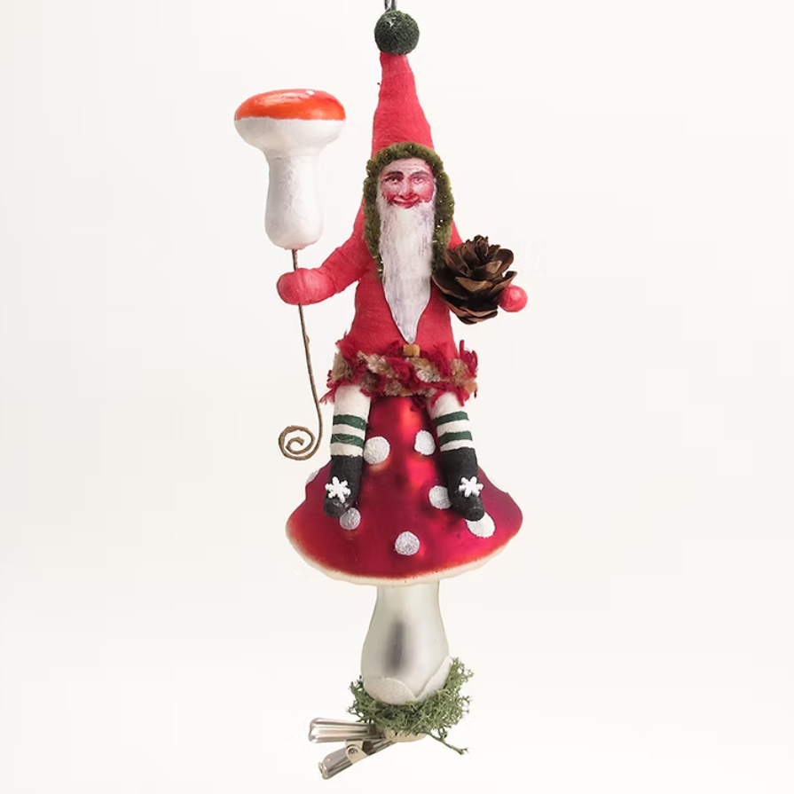 A whimsical Christmas ornament depicting a gnome sitting on a large red and white mushroom, holding a small pinecone and a string, set against a plain white background.