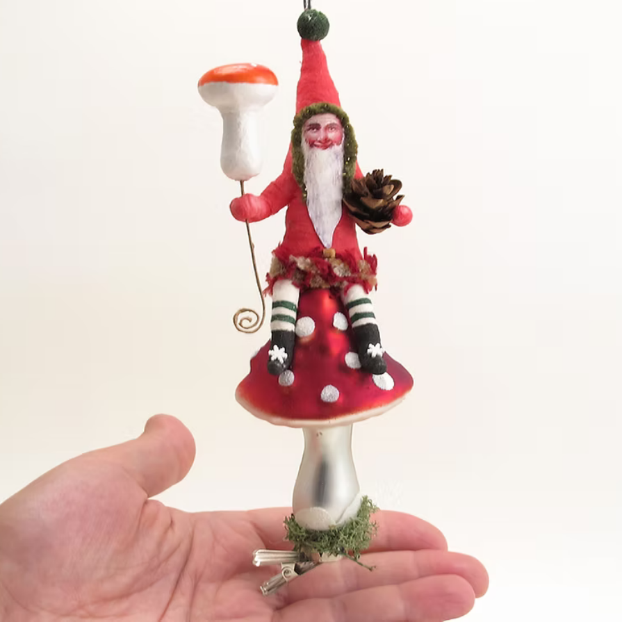A hand holds a small Santa Claus ornament perched on a red-and-white mushroom cap. The ornament features a red hat and coat, white beard, black-and-white striped legs, and holds a white mushroom and a pinecone, against a plain light background.