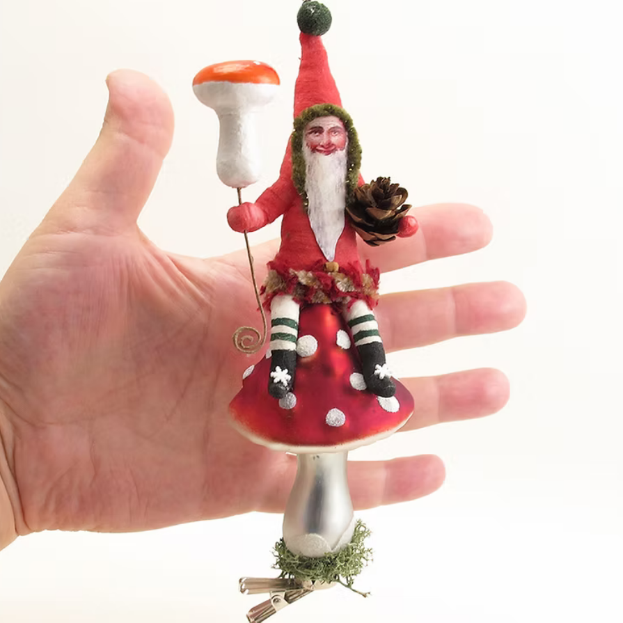 A hand holding a small decorative ornament shaped like a fairy sitting on a red mushroom cap, with a white mushroom topper and silver stem, against a plain white background.