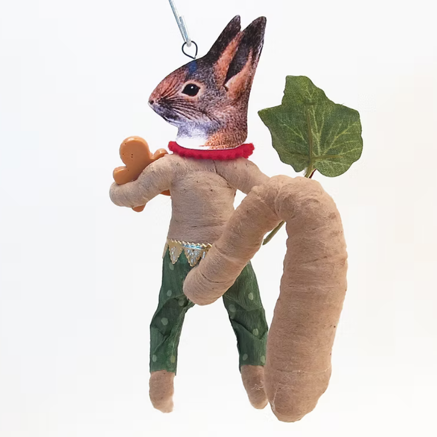 A whimsical handmade rabbit ornament with a realistic rabbit head, long tan body, green patterned pants, a red collar, and a large green leaf on its back, shown against a plain white background.