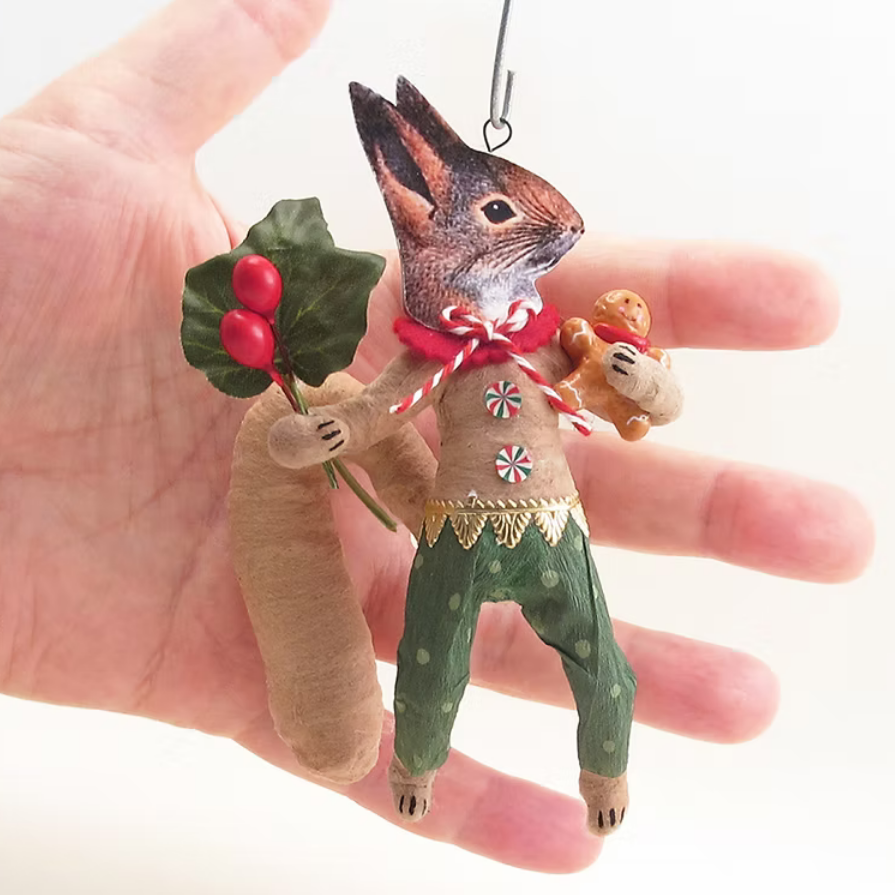 A hand holding a whimsical rabbit-shaped ornament or figurine with a brown fur-textured head, green pants, a red-and-white striped neckerchief, gold trim, and a small teddy bear in one paw, against a plain white background.