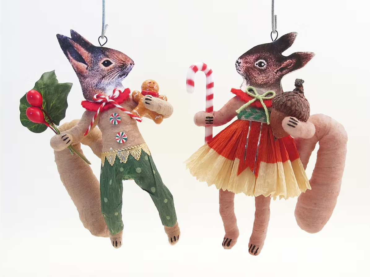 Two handmade felt rabbit ornaments hanging against a plain white background. One rabbit is dressed in green pants with a red bow and holds a sprig of holly and a gingerbread cookie.