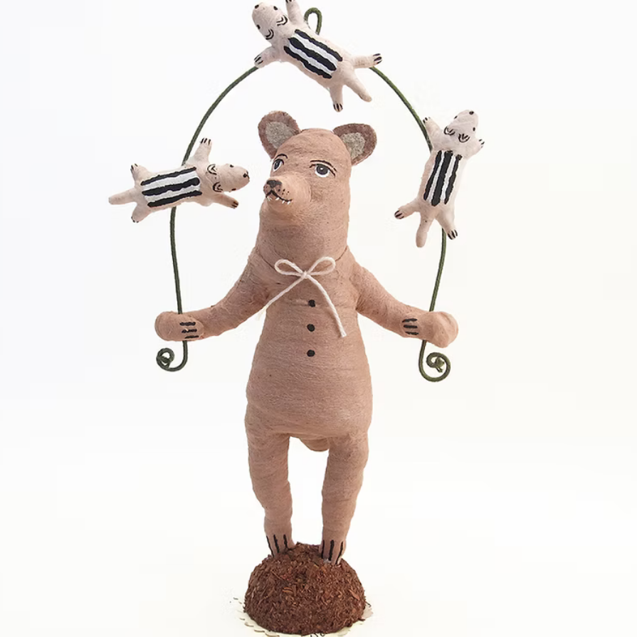A handmade fabric pig figurine standing on a small brown base, holding a metal skipping rope with four small striped pigs jumping around it, photographed against a plain white background.