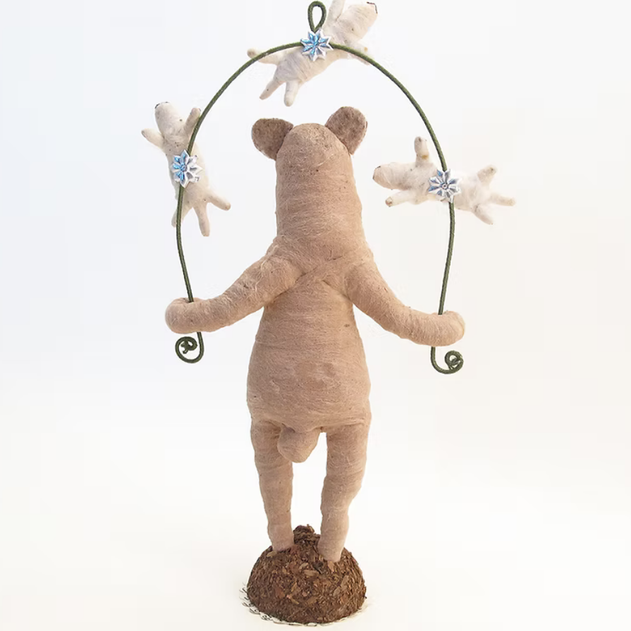 A handmade beige fabric bear figurine standing on a brown textured base, holding a green metal hoop with small white shells and blue floral decorations, photographed against a seamless white background.