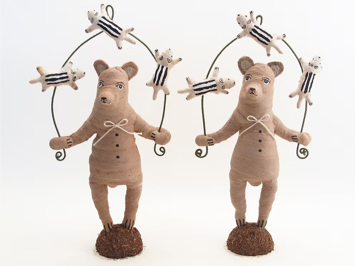 Two handmade clay bear figurines holding wire hoops with small striped pig dolls, standing on brown textured bases against a white background.