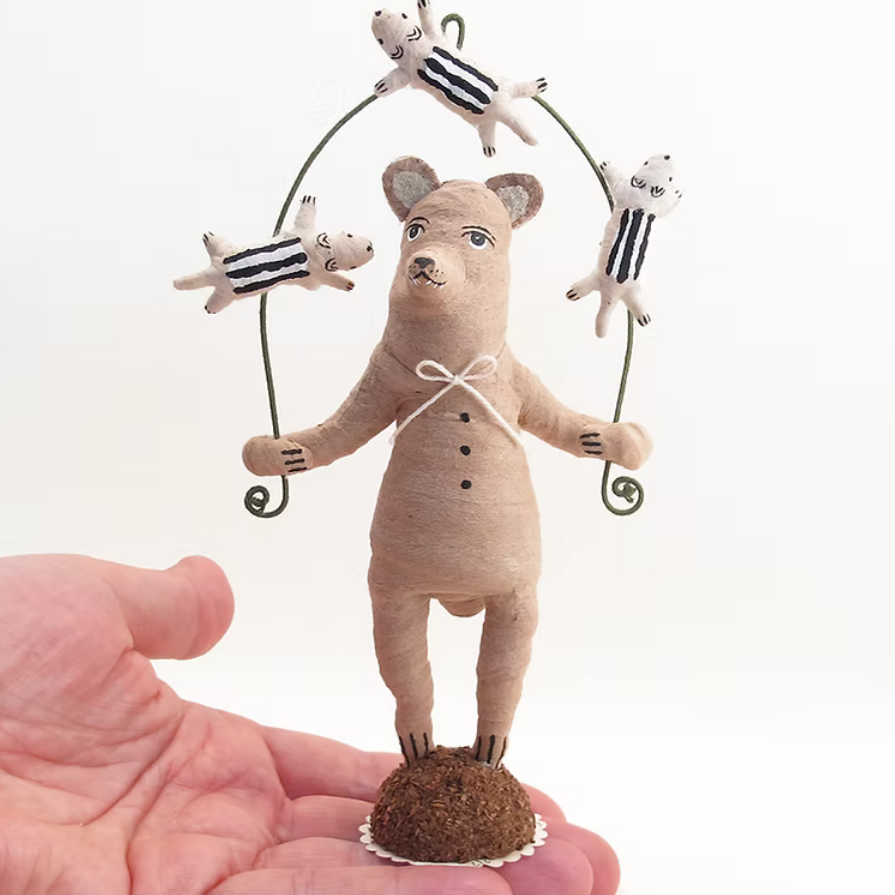 A miniature handmade doll of a bear balancing a wire merry-go-round with four striped sheep, held in a person's hand against a white background.