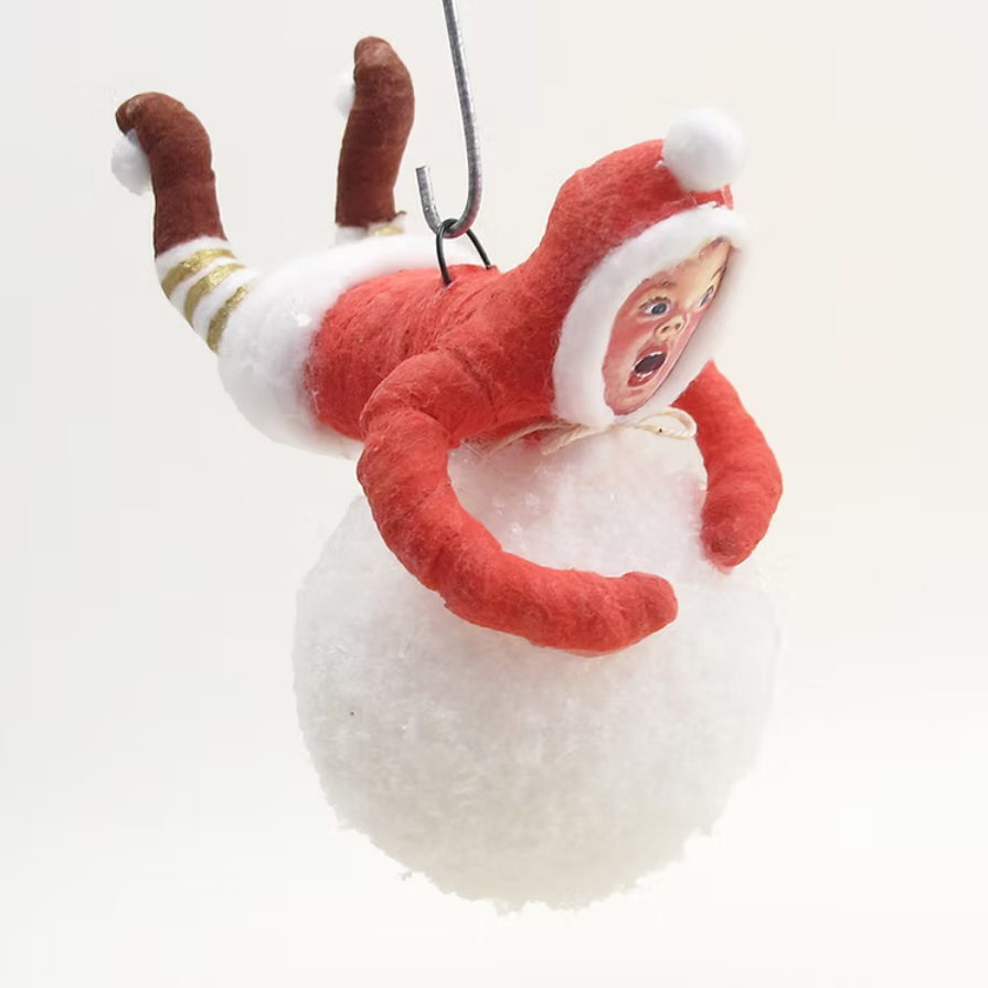 A novelty Christmas ornament featuring a small doll dressed in a red Santa-style suit with white trim, crawling on a white fluffy ball, suspended by a wire hook against a plain white background.