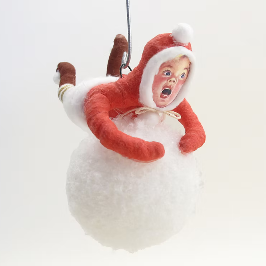 A novelty Christmas ornament shaped like a baby doll in a red and white Santa-style hood lying on top of a large white snowball, suspended against a plain light background.