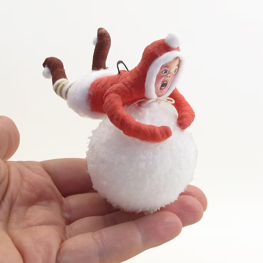 A small handmade plush ornament shaped like a person in a red Santa-style suit riding on the back of a white fluffy ball, held in a person's hand against a plain white background.