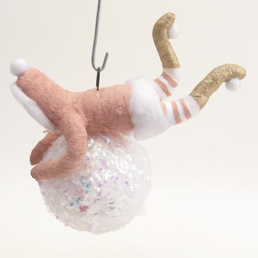 A handmade felt cat hanging ornament shown upside down from a metal hook. The felt cat has a pink body, white paws and tail tip, and gold-striped legs, hanging above a clear bubble ball filled with glitter.