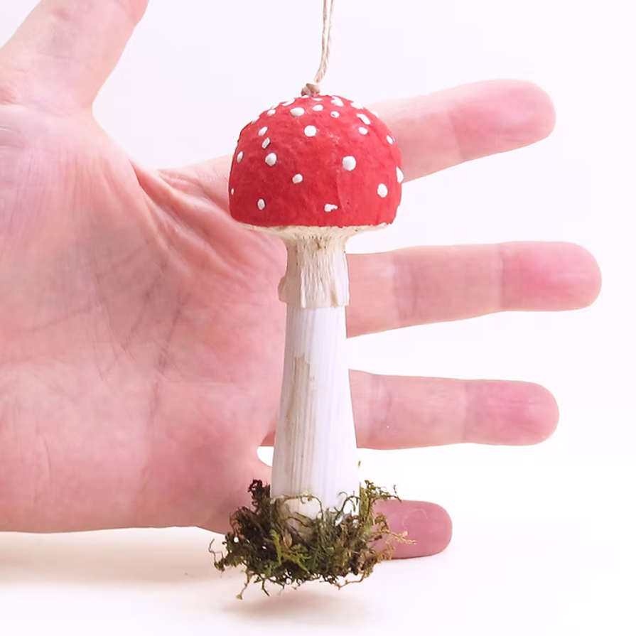 A small decorative mushroom ornament with a red cap, white spots, a white stem, and green moss at the base, held in a person's hand against a plain white background.