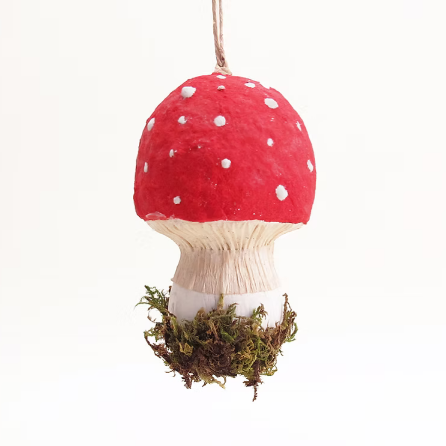 A hanging mushroom ornament with a red cap, white spots, a white stem, and green-brown moss at the base, suspended against a plain white background.