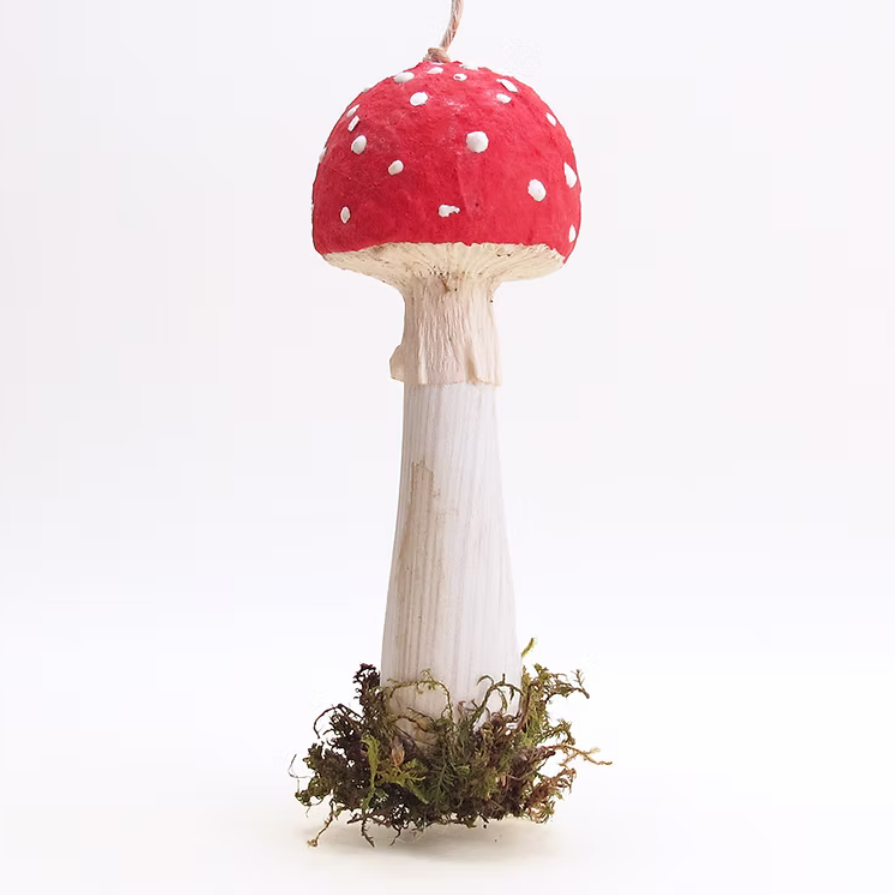 A decorative hanging ornament shaped like a mushroom, featuring a red cap with white spots, a white stem, and a base wrapped in brown and green moss, suspended against a plain white background.